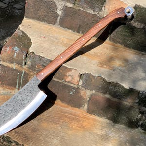 May include: A large cleaver with a dark wooden handle and a wide, textured blade. The handle is curved and has small rivets. A metal ring is attached at the end of the handle. The blade has a polished edge. The cleaver is designed for chopping.