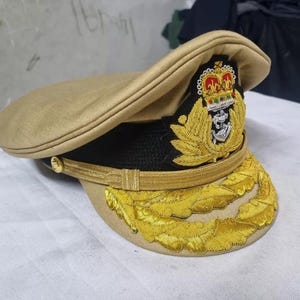 May include: A tan military style hat with a gold braid band and a gold embroidered emblem featuring a crown and an anchor. The hat has a black band with a gold trim.
