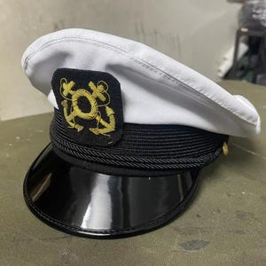 May include: A white and black sailor hat with a gold embroidered emblem featuring a circle and anchors. The hat has a black brim and a black band with a rope design.
