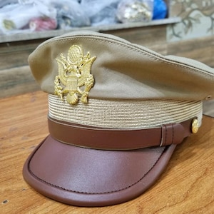 Op de afbeelding: Een beige militaire hoed met een bruine leren rand en een gouden adelaarsembleem aan de voorkant. De hoed heeft een gouden gevlochten rand rond de kroon.