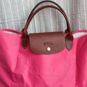May include: A bright pink tote bag with brown leather accents. The bag features brown leather handles and a flap closure with a gold-colored button. The flap is embossed with a logo. The bag is made of a durable, lightweight material.