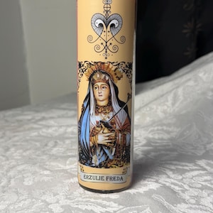May include: A cylindrical candle with a cream-coloured label featuring an illustration of a woman wearing a crown and holding a cross. The label also includes the text "ERZULIE FREDA" and decorative symbols.