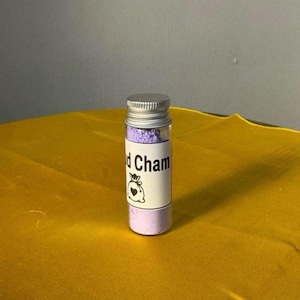 May include: A small glass vial with a silver cap, filled with a light purple powder. The vial has a white label with the text "d Cham" and a small graphic of a bag with a heart. The vial is set against a yellow background.