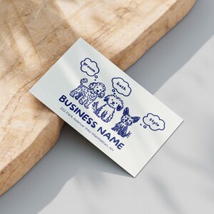 This image shows a realistic mockup of the dog grooming business card placed on a light wooden surface. The card features navy line-art illustrations of three dogs with “groom,” “bath,” and “style” speech bubbles, plus space for the business name and address. The soft shadows and warm texture make the card appear printed and professionally photographed. Ideal for showcasing the final look of this editable Canva grooming business card template.