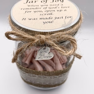 May include: A clear glass jar with a metal lid, filled with pink scrolls. The lid has text that reads "Jar of Joy." The jar is decorated with twine, lace, and a heart-shaped charm that says "Love."