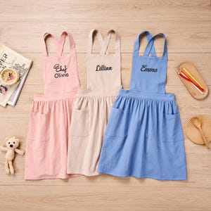 Puede incluir: Tres delantales personalizados en rosa, beige y azul. El delantal rosa tiene bordado "Chef Olivia", el delantal beige "Lillian" y el delantal azul "Emma". Cada delantal tiene un bolsillo y una falda fruncida.
