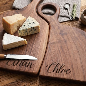 Könnte beinhalten: Zwei Holz-Käsebretter mit Griffen, eines mit dem Namen "Anan" und das andere mit "Chloe" graviert. Sie zeigen verschiedene Käsesorten und ein kleines Messer. Das Holz hat eine tiefe, dunkelbraune Farbe.