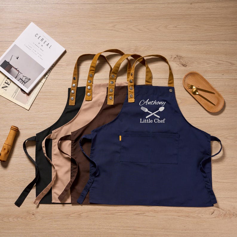 Op de afbeelding: Vier schorten in zwart, beige, bruin en marineblauw met leren bandjes. Het marineblauwe schort heeft de tekst "Anthony Little Chef" met een gekruist vork- en lepelontwerp. De schorten liggen op een houten oppervlak.