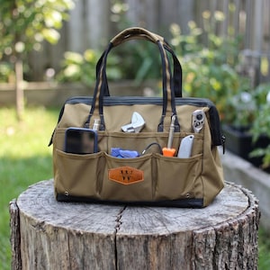 Könnte beinhalten: Eine braune Werkzeugtasche aus Canvas mit schwarzen Akzenten, gefüllt mit Werkzeugen und einem Smartphone. Die Tasche hat ein Lederetikett mit den Initialen "VV". Die Tasche steht auf einem Baumstumpf.