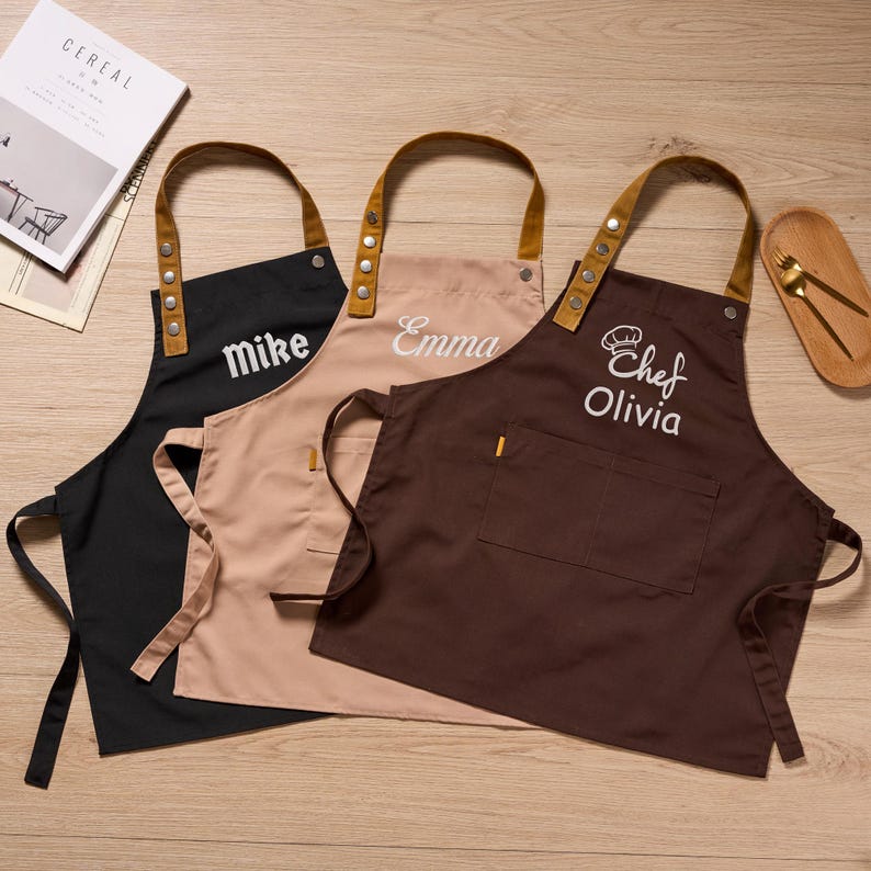 Op de afbeelding: Drie gepersonaliseerde schorten in zwart, beige en bruin, elk geborduurd met een andere naam in wit. De schorten hebben bruine leren bandjes en zakken. E&eacute;n schort heeft de naam "Mike", een andere "Emma" en de derde "Chef Olivia".