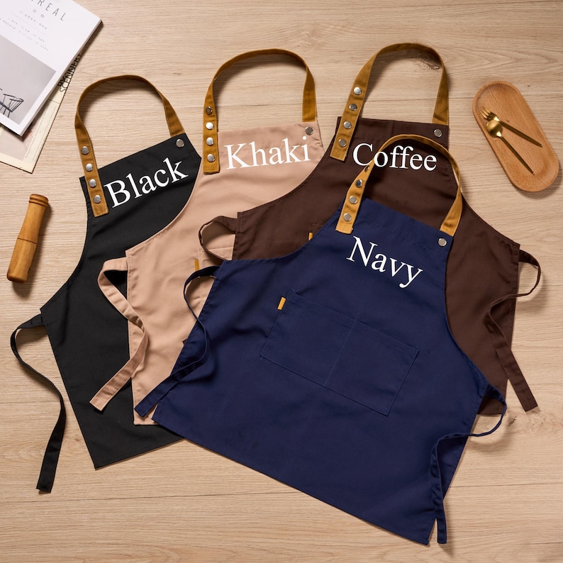 Op de afbeelding: Vier schorten in zwart, kaki, koffie en marineblauw, elk met een contrasterende bruinleren nekriem en een zak. De schorten hebben de kleurnaam in het wit erop gedrukt.