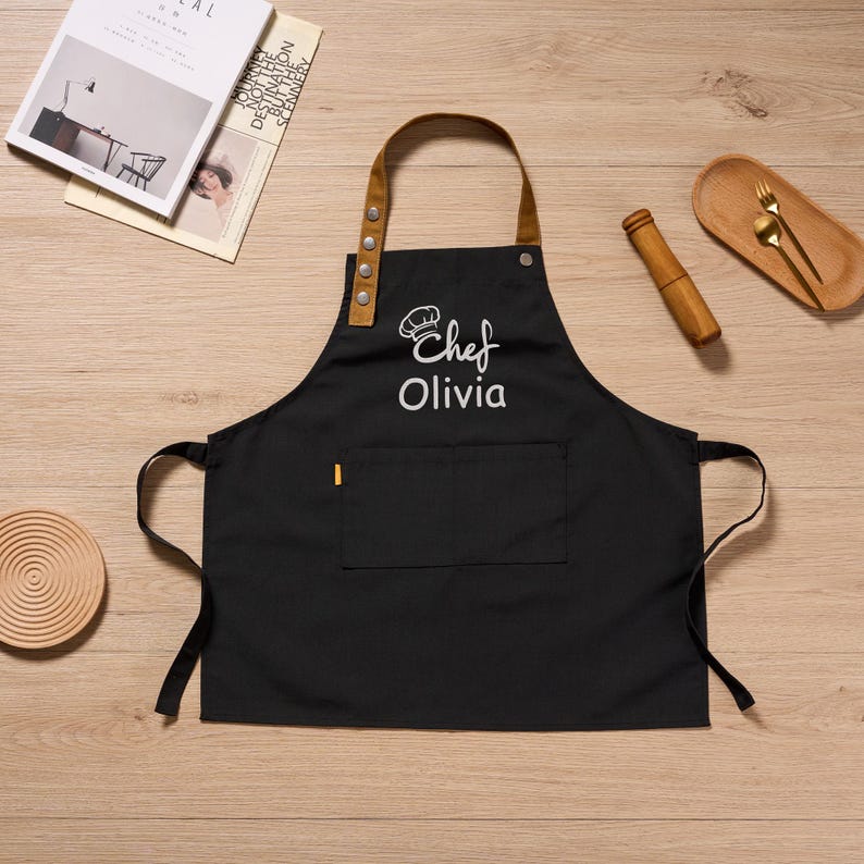 Op de afbeelding: Zwarte schort met een bruine leren nekband en zak. De schort heeft de tekst "Chef Olivia" in het wit geborduurd. Een kleine gele accent is op de zak. De schort ligt op een houten oppervlak.