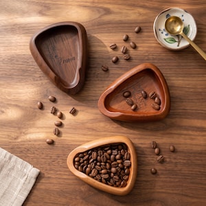 May include: Three triangular wooden bowls with rounded corners. One bowl is engraved with the name Emma, another with the name Jean, and the third is filled with roasted coffee beans. A small ceramic dish with a gold spoon sits nearby.