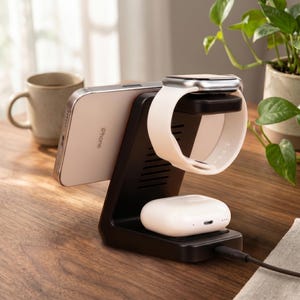 Op de afbeelding: Een zwart oplaadstation met een zilveren iPhone, een witte Apple Watch met een witte band en witte oortelefoons. Het station staat op een houten oppervlak, met een koffiemok en een plant op de achtergrond.