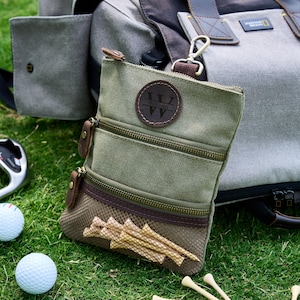 May include: A green canvas golf accessory bag with multiple zipped compartments and a brown leather patch with the name "William". The bag is filled with golf tees and is next to golf balls and a golf club.
