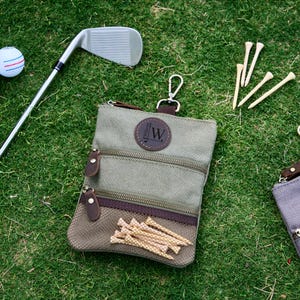 Op de afbeelding: Een golftas met meerdere vakken met ritssluiting, een bruin leren patch met de initialen "JW" en een verzameling golftees. Een golfclub en een golfbal zijn ook zichtbaar op het groene gras.