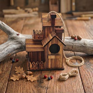 May include: A handcrafted wooden birdhouse with a chimney, balcony, and round entry hole. The birdhouse is made of various shades of brown wood and has a rope for hanging. Acorns and oak leaves are scattered around the base, suggesting a rustic theme.