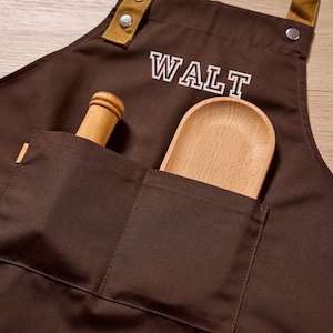 Op de afbeelding: Een bruine schort met de naam "WALT" in witte blokletters. De schort heeft een zak met een houten lepel en een houten deegroller. De schort heeft bruine en bruin/beige accenten.