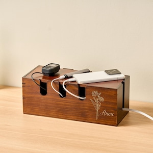 May include: A wooden cable organizer in a rich brown finish. It features multiple slots for cords and a top surface for devices. The organizer has a floral design and the name "Anna" engraved on the side. A smartwatch, a pen, and a smartphone are on top.