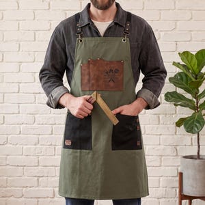 May include: An olive green apron with black pockets and a brown leather chest pocket featuring a graphic of a flame and spatula. The apron has adjustable black straps with metal hardware and is held with a brass square tool in the center.