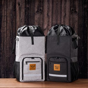 May include: Two insulated cooler backpacks, one light grey and one dark grey, with black detailing. Each has a front pocket, side mesh pocket, and a reflective stripe. The light grey backpack has the name "Andrew" on a leather patch.