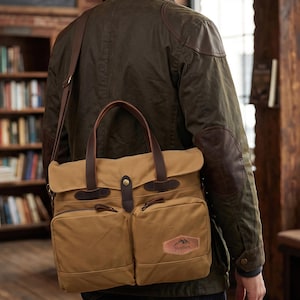 Op de afbeelding: Een beige canvas schoudertas met twee voorzakken, donkerbruine lederen handvatten en een schouderriem. De tas heeft een klepsluiting met een koperen drukknoop en een lederen patch met de tekst Stephen en een icoon van een berg.