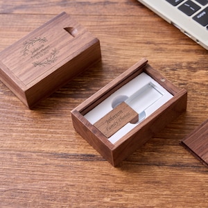 以下が含まれることがあります： 木製のUSBフラッシュドライブとボックスのセット。ボックスは開いており、木製の端が付いた透明なUSBドライブが表示されています。ボックスとUSBの端には「Johnson Family Photos」と日付05-18-2023が刻印されています。セットは木製の表面にあります。