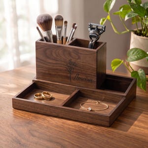 May include: A dark brown wooden vanity organizer featuring a tall compartment for makeup brushes and a razor, and a lower tray with two sections. The front is engraved with a floral design and the name Ameira. Two gold rings and a gold chain bracelet rest inside.