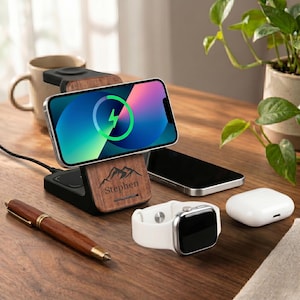 Op de afbeelding: Een houten oplaadstation met telefoon, horloge en oortelefoons. De telefoon toont een oplaadsymbool. Het station heeft een bergontwerp en de naam "Stephen" gegraveerd. Een pen en een mok zijn ook zichtbaar.