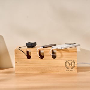 Könnte beinhalten: Eine Kabelbox aus Naturholz mit drei Schlitzen für Kabel. Ein schwarzes Gerät, ein silberner Stift und ein weißes Smartphone sind angeschlossen. Die Box hat ein rundes Emblem mit dem Buchstaben "M" und dem Namen "MICHAEL".