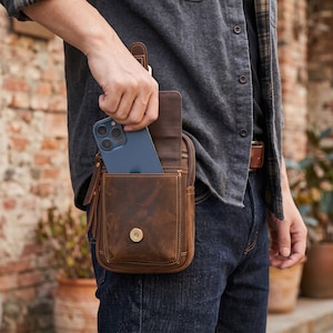 May include: A brown leather belt pouch featuring a front pocket with a gold-toned magnetic snap closure and a main compartment containing a dark blue smartphone. The bag has visible stitching and leather zipper pulls attached to the side hardware.