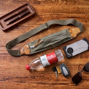 May include: An olive green canvas fanny pack with a tan leather accent featuring the initials CM, a matching olive green adjustable strap, and a silver zipper. The bag is accompanied by two pens in a wooden tray, a smartphone, a water bottle, keys, and sunglasses.