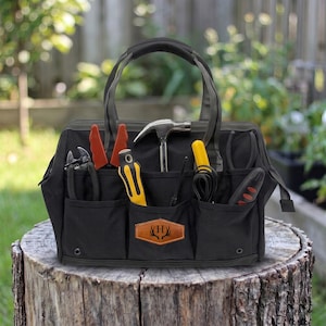 May include: Black canvas tool bag with multiple pockets, containing tools such as pliers, a hammer, and screwdrivers. The bag features a leather patch with a deer antler design and the letter "H". The bag is placed on a tree stump.