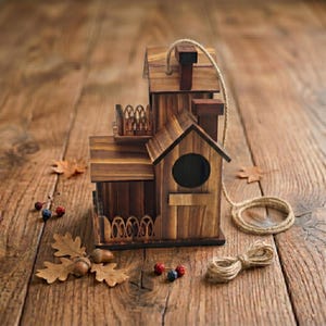May include: A multi-level wooden birdhouse with a chimney, round entry hole, and decorative railing. The birdhouse is made of stained wood and has a rope for hanging. Acorns and leaves are scattered around the birdhouse.