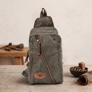 May include: An olive green canvas sling backpack featuring a diagonal zippered front pocket with a brown leather pull tab and a matching leather patch at the bottom displaying a black logo with the letter W and horizontal lines.