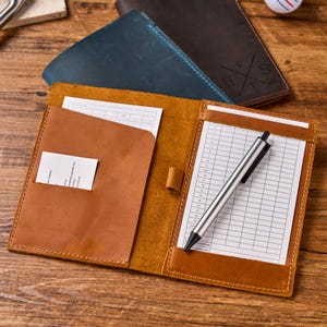 May include: A tan leather golf score card holder with a pen and score card. The holder is open, revealing a white score card and a small pocket. Other leather items are in the background.