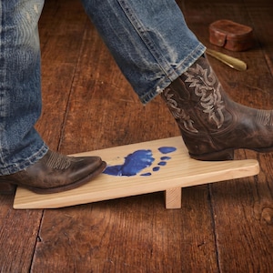 May include: A light-colored wooden boot jack with a blue footprint design. The boot jack is on a wooden surface, with a person's boots on the jack. The boots are brown leather cowboy boots.