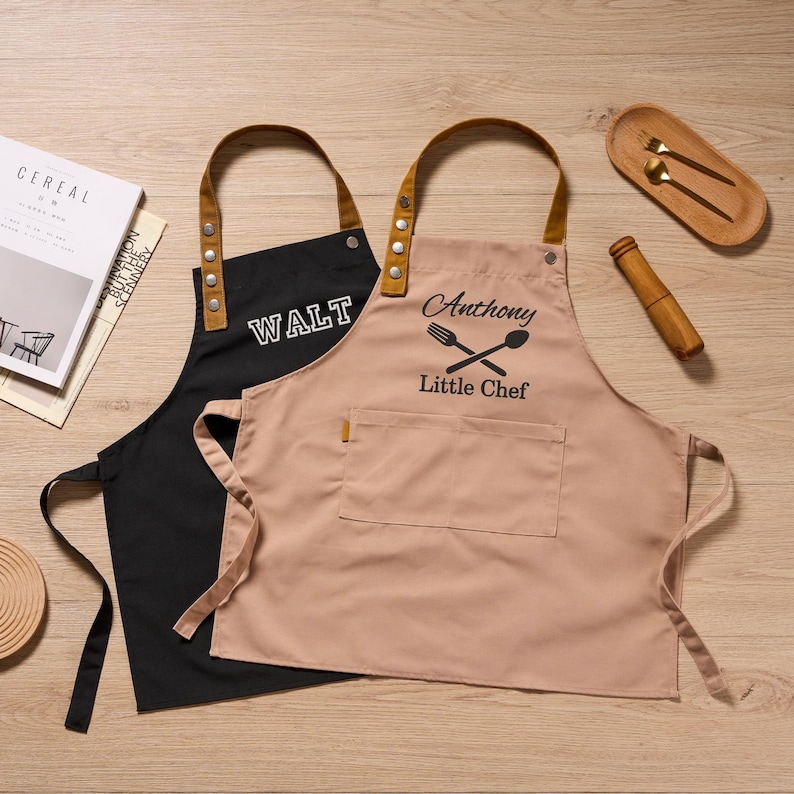 Op de afbeelding: Twee schorten, &eacute;&eacute;n zwart en &eacute;&eacute;n bruin, met bruine leren bandjes en de tekst "WALT" en "Anthony Little Chef". De schorten hebben zakken en liggen op een houten oppervlak. Een houten lepel- en vorkset en een deegroller zijn ook zichtbaar.