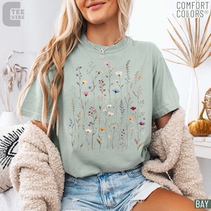 May include: A light green Comfort Colors t-shirt featuring a watercolor floral design. The design includes various wildflowers in shades of pink, purple, yellow, and blue. The shirt also has the text "TEE VIBE" on the upper left.