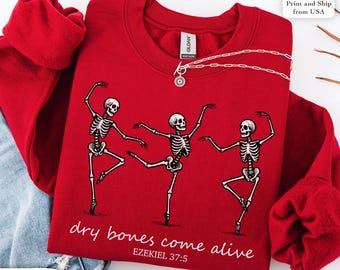 May include: A red sweatshirt featuring a graphic of three dancing skeletons. The phrase "dry bones come alive" and the Bible verse Ezekiel 37:5 are printed below the skeletons. A silver necklace with a pendant is draped on the sweatshirt.