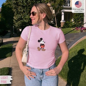 May include: Light pink t-shirt featuring a classic Mickey Mouse graphic. The shirt has a crew neck and short sleeves. The text "LIGHT PINK" is in a banner at the bottom left.