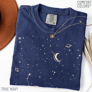 Op de afbeelding: Een marineblauw Comfort Colors T-shirt met een hemels ontwerp met sterren, planeten en een halve maan. Een gouden ketting met een hanger ligt op het shirt. De woorden "TRUE NAVY" staan onderaan.