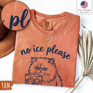 Puede incluir: Camiseta naranja Comfort Colors con la frase "no ice please" y un gr&aacute;fico de gato. El gato sostiene un vaso de hielo. Un collar dorado con un colgante est&aacute; en la camisa.