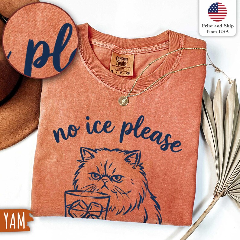Puede incluir: Camiseta naranja Comfort Colors con la frase "no ice please" y un gr&aacute;fico de gato. El gato sostiene un vaso de hielo. Un collar dorado con un colgante est&aacute; en la camisa.
