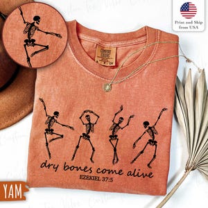 May include: A coral-colored t-shirt featuring a graphic of dancing skeletons. The phrase "dry bones come alive" and the Bible verse Ezekiel 37:5 are printed below the skeletons. A gold necklace with a pendant is draped on the shirt.