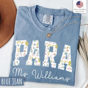 Pode incluir: Uma camiseta azul jeans Comfort Colors com a palavra "PARA" em letras brancas preenchidas com um padrão floral. O nome "Ms. Williams" está escrito abaixo em itálico. Um colar de ouro repousa sobre a camisa.