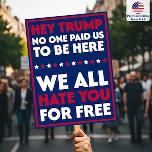 Op de afbeelding: Een blauw rechthoekig bord met witte en rode tekst, vastgehouden door een persoon in een menigte. De tekst luidt: "HEY TRUMP NO ONE PAID US TO BE HERE WE ALL HATE YOU FOR FREE". Er is een rij rode en witte sterren.