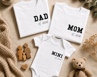 Puede incluir: Camisetas blancas y un body de bebé con texto negro que dice "DAD est. 2026", "MOM est. 2026" y "MINI est. 2026". También hay una figura de familia de madera, un corazón y un osito de peluche. Los artículos están colocados sobre una superficie neutra.