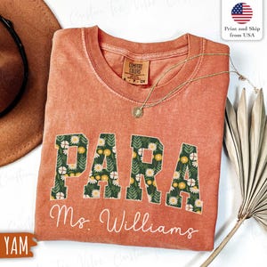 Pode incluir: Uma camiseta cor de terracota com a palavra "PARA" em um design floral. Abaixo da palavra "PARA" está o nome "Ms. Williams" em itálico. Um colar de ouro com um pingente está drapeado na camisa.