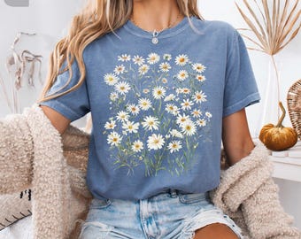 以下が含まれることがあります： デイジーの花柄が特徴のブルージーンズカラーのTシャツ。半袖とクルーネックです。「TEE VIBE」の文字が左上に、「COMFORT COLORS」の文字が右上にあります。「BLUE JEAN」の文字が下にあります。